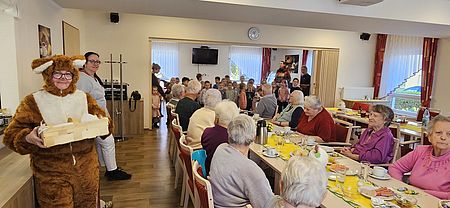 Der Osterhase besucht die Tagespflege in Osterweddingen Eine Tafel an der Senioren sitzen, dahinter eine Gruppe Kinder. Vorne links eine Mitarbeiterin im Osterhasenkostüm.