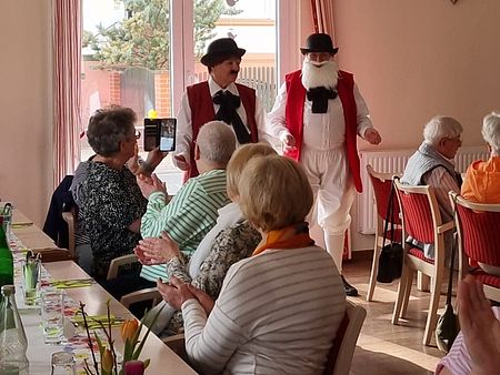 Die "Wildecker Herzbuben" (Double) in Eilsleben zu Besuch Ein Wildecker Herzbuben Double steh vor einem Fenster. Senioren an einer Tafel klatschen.