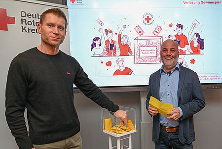 Auslosung des Sudoku-Gewinnspiels durch Martin Tunsch und Torsten Winkelmann Zwei Männer stehen neben eine gläsernen Würfel mit Losen
