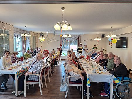 Fasching in der Tagespflege in Eilsleben mehrere Seniorinnen und Senioren sitzen an zwei langen Tafeln, die nach Faschingsart geschmückt sind