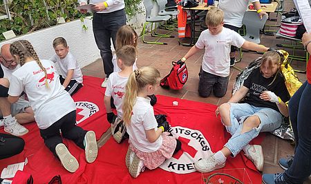 Kinder und Jugendliche treten im gemeinsamen Wettbewerb an mehrere Kinder hocken auf roten Decken und üben die Behandlung von Verletzten