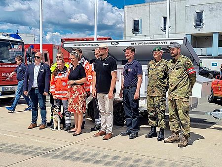 Übergabe des Hunde-Anhängers im Rahmen einer Übung Mehrere Personen, unter Anderem 2 Bundeswehrsoldaten und zwei Sanitäter stehen vor einem großen Anhänger für Hunde